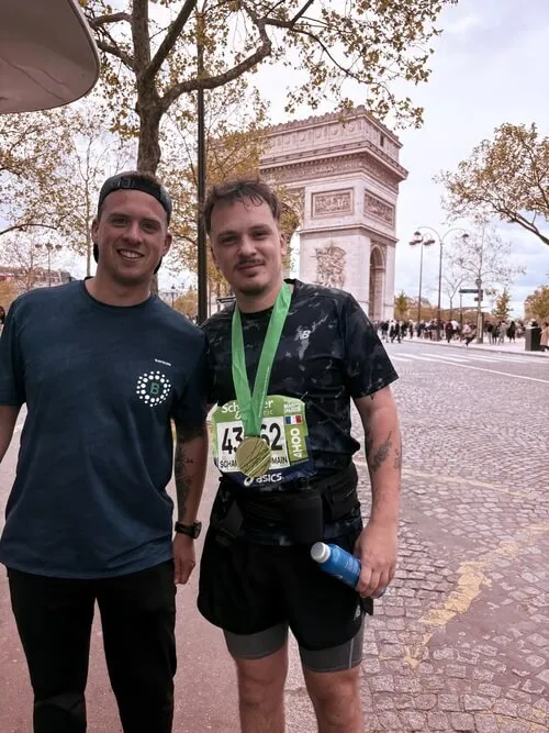 Deux coureurs souriants après une course, médaille autour du cou, devant l'Arc de Triomphe à Paris